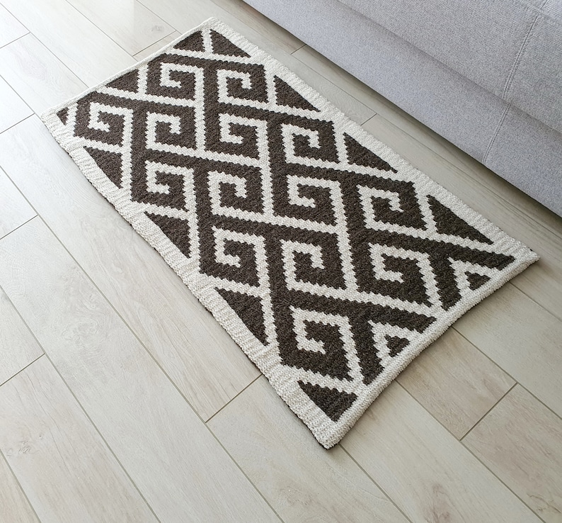 May include: A brown and white geometric patterned rug with a white border. The rug is made of a soft, woven material.