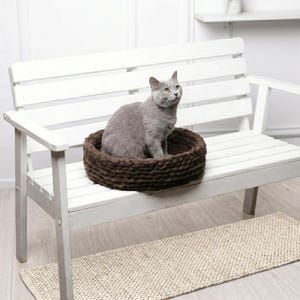 A gray short-haired cat sitting upright inside a round dark brown chunky knit wool pet bed, placed on a white wooden bench in a bright minimalist interior with a light hardwood floor and a woven jute rug underneath.