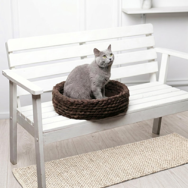 A gray short-haired cat sitting upright inside a round dark brown chunky knit wool pet bed, placed on a white wooden bench in a bright minimalist interior with a light hardwood floor and a woven jute rug underneath.