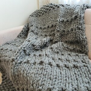 A chunky knit blanket in a soft gray color is draped over a light-colored sofa. The blanket features a textured pattern with raised, rounded elements. The thick yarn creates a cozy and inviting appearance, perfect for home decor.