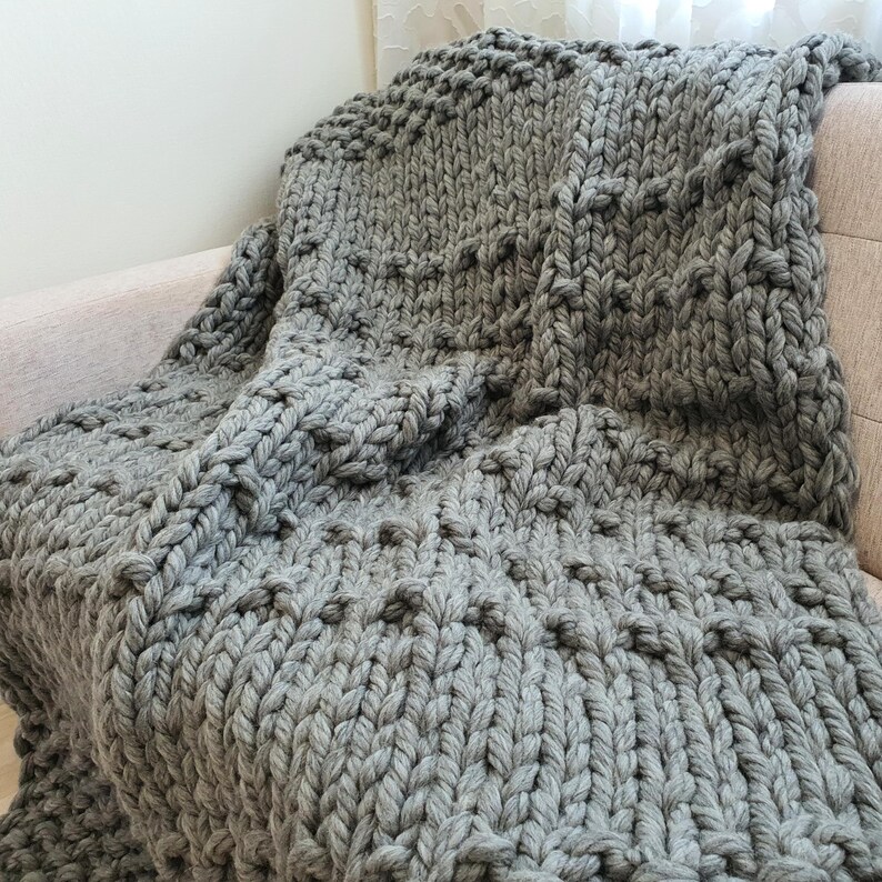 A chunky knit blanket in a soft gray color is draped over a light-colored sofa. The blanket features a textured pattern with raised, rounded elements. The thick yarn creates a cozy and inviting appearance, perfect for home decor.