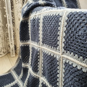 May include: A crocheted throw blanket with a square pattern in shades of blue and gray. The blanket is made with a chunky yarn and has a textured, hand-stitched look.