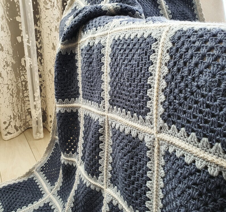 May include: A crocheted throw blanket with a square pattern in shades of blue and gray. The blanket is made with a chunky yarn and has a textured, hand-stitched look.