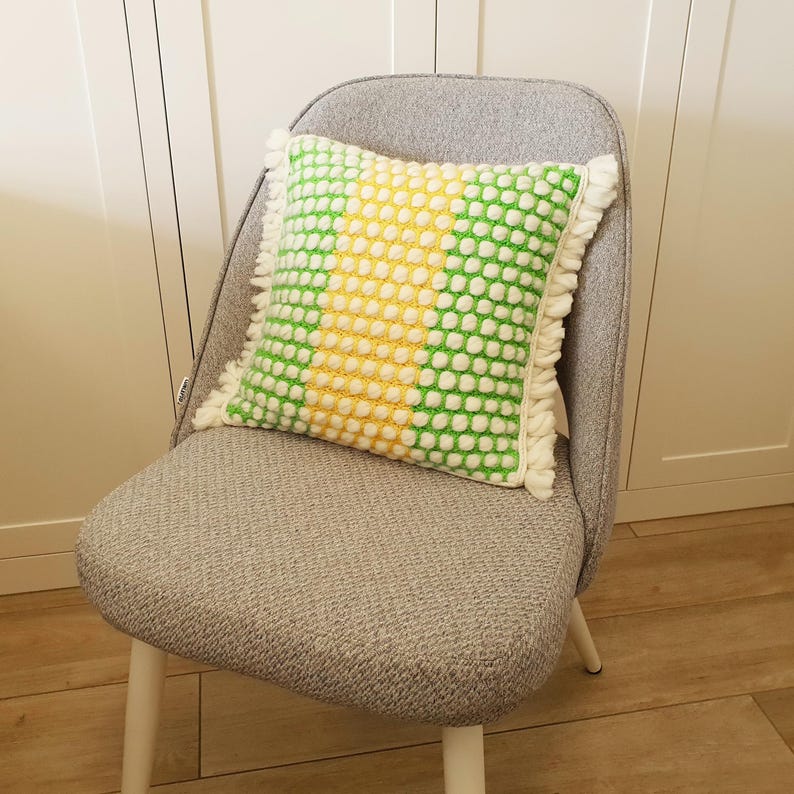 May include: A decorative square pillow with a textured pattern in green, yellow, and white, with a fringe trim. The pillow is placed on a gray upholstered chair with white legs. The pillow's design features a grid of white dots on a green and yellow background.