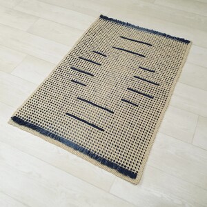 May include: A beige and blue woven rug with a fringe border. The rug has a geometric pattern of dark blue lines.