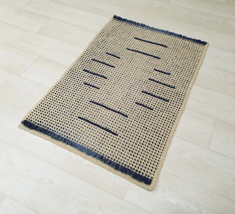 May include: A beige and blue woven rug with a fringe border. The rug has a geometric pattern of dark blue lines.