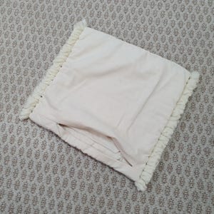 May include: Cream-colored pillow with a textured, fringed border. The pillow has a soft, folded appearance and is made of a light-colored fabric. The fringing adds a decorative element to the pillow's edge.