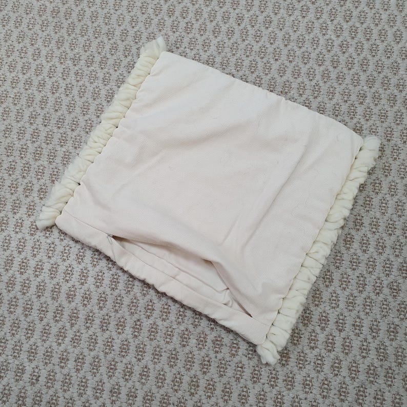 May include: Cream-colored pillow with a textured, fringed border. The pillow has a soft, folded appearance and is made of a light-colored fabric. The fringing adds a decorative element to the pillow's edge.