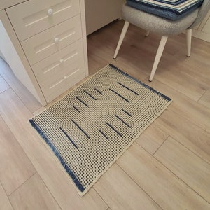 May include: A beige and blue woven rug with a fringe border. The rug has a geometric pattern of thin blue lines on a beige background.