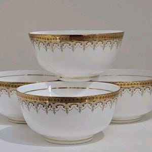 Könnte beinhalten: Vier weiße Keramikschalen mit goldenen Rändern und dekorativen Goldmustern. Die Schalen sind gestapelt und angeordnet, um die komplizierten Details des Golddesigns zu zeigen. Die Schalen sind ein klassisches Design.