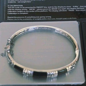 May include: A silver-toned hinged bangle bracelet featuring alternating smooth black segments and textured silver metal sections. The bracelet includes a secure clasp mechanism with a small attached safety chain, resting on a digital scale.