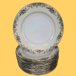 Puede incluir: Una pila de platos de porcelana blanca con un diseño de borde floral y azul. Los platos tienen un borde dorado y un delicado patrón de flores rosas, amarillas y moradas. Los platos están apilados uno encima del otro.