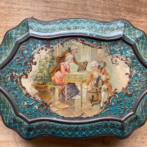 Puede incluir: Una lata vintage de metal azul verdoso con un borde elevado y adornado. La lata presenta una ilustración colorida de un grupo de personas tocando música en una habitación. La escena incluye una mujer tocando el piano, un hombre tocando el violonchelo y otra mujer escuchando.