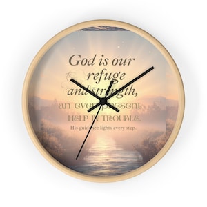 Op de afbeelding: Een ronde wandklok met een licht houten frame. De wijzerplaat toont een serene landschap met een rivier en de tekst "God is onze toevlucht en kracht, een altijd aanwezige hulp in de nood." Zwarte wijzers geven de tijd aan.