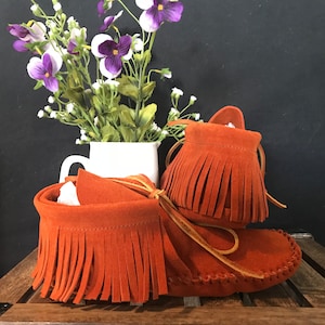 May include: A pair of orange suede moccasins with fringe. The moccasins are on a wooden surface.