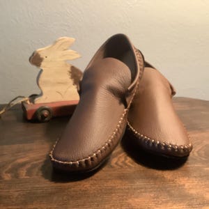 May include: A pair of brown leather moccasins with a decorative stitch around the edge.