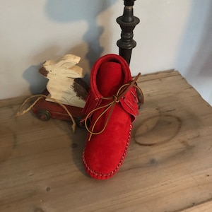 May include: A pair of red leather moccasins with brown leather laces and fringe. The moccasins are sitting on a wooden surface.