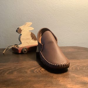 May include: A brown leather moccasin with a stitched edge. The moccasin is on a wooden surface next to a wooden toy rabbit.