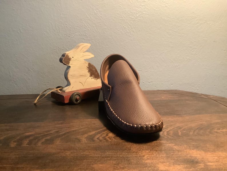 May include: A brown leather moccasin with a stitched edge. The moccasin is on a wooden surface next to a wooden toy rabbit.