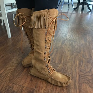 May include: Tan suede knee-high boots with fringe detail at the top. The boots feature a lace-up design and are adorned with small, round studs along the sides. The soles have a moccasin-style construction.