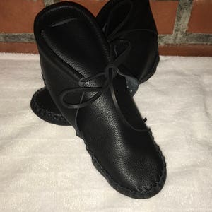 May include: A pair of black leather moccasins with a decorative stitch around the sole. The moccasins have a lace-up closure and are made of soft, supple leather.