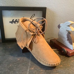May include: A pair of brown leather moccasins with fringe and laces. The moccasins are tied with a brown leather lace and are sitting on a wooden surface.