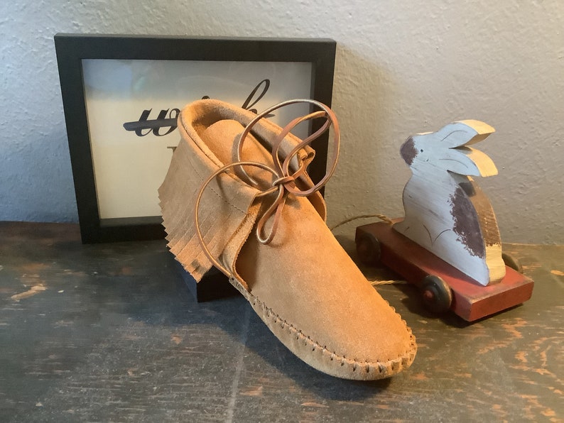 May include: A pair of brown leather moccasins with fringe and laces. The moccasins are tied with a brown leather lace and are sitting on a wooden surface.