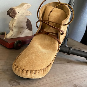 Pu&ograve; includere: Stivali mocassino in camoscio beige con lacci marroni. Gli stivali hanno una cucitura cucita attorno al bordo della suola.