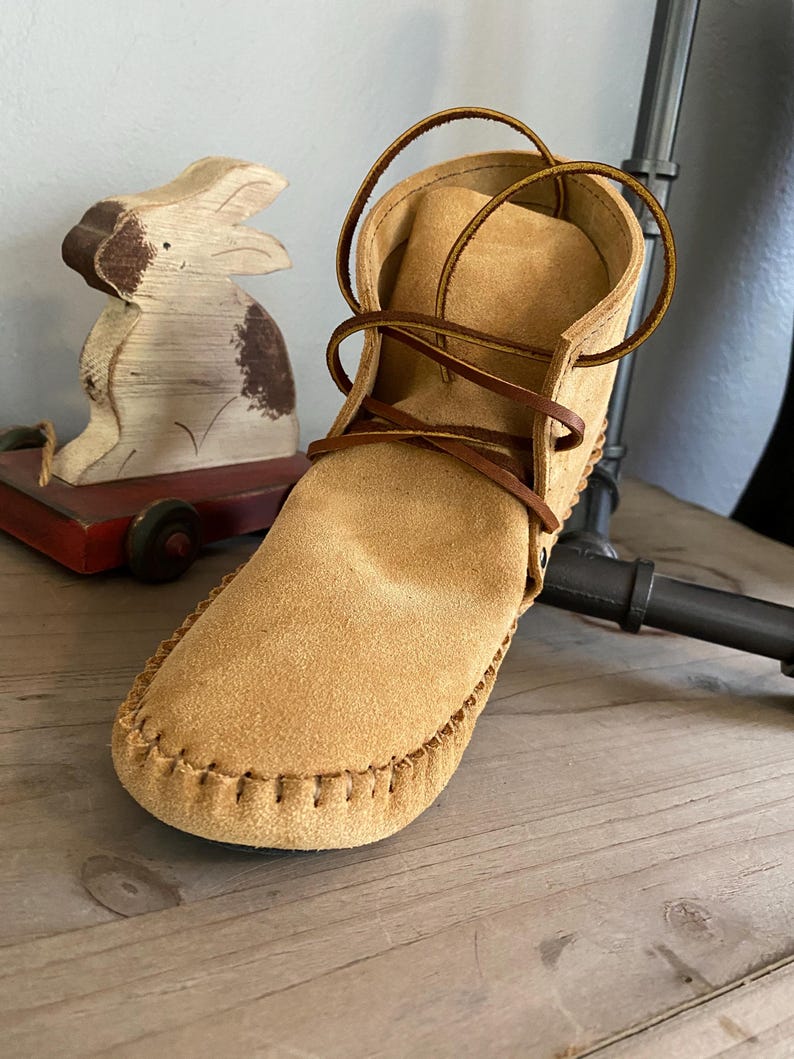 Pu&ograve; includere: Stivali mocassino in camoscio beige con lacci marroni. Gli stivali hanno una cucitura cucita attorno al bordo della suola.
