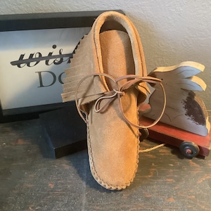 May include: A brown leather moccasin with fringe and a leather lace. The moccasin is sitting on a wooden surface.