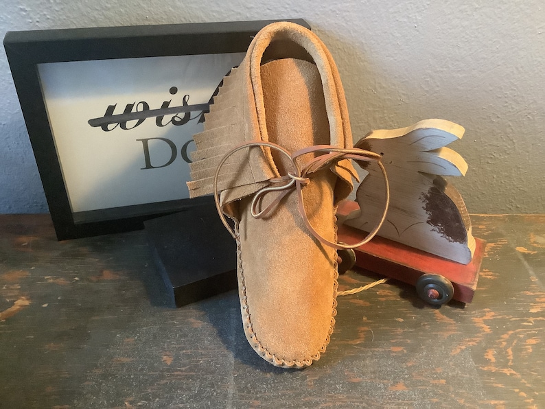 May include: A brown leather moccasin with fringe and a leather lace. The moccasin is sitting on a wooden surface.