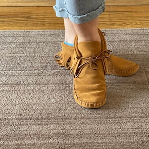 May include: A pair of brown suede moccasins with fringe detail. The moccasins have a lace-up closure and are worn on a person's feet.