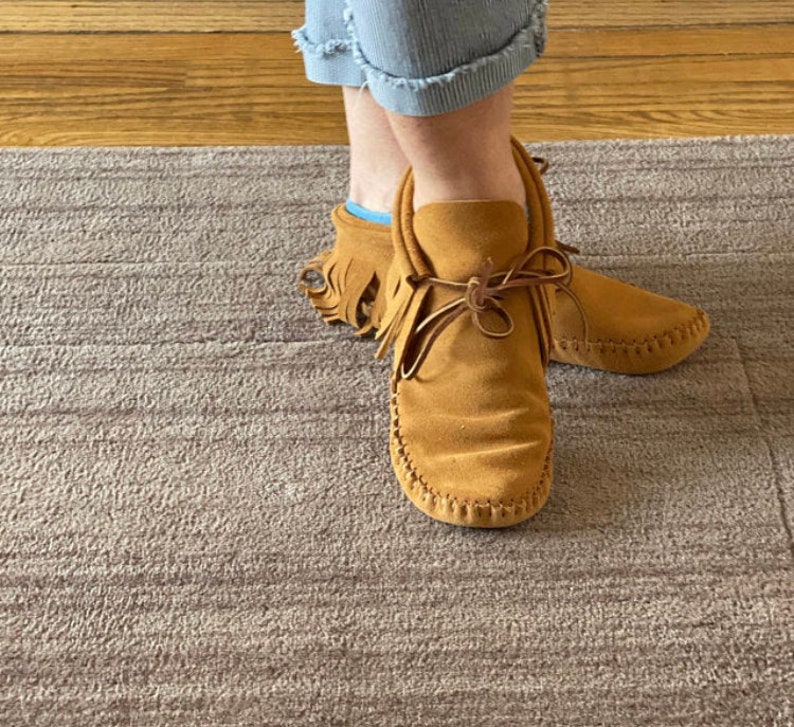 May include: A pair of brown suede moccasins with fringe detail. The moccasins have a lace-up closure and are worn on a person's feet.