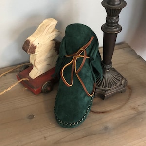 May include: A pair of dark green suede moccasins with brown leather laces and fringe. The moccasins are tied with a bow and are sitting on a wooden surface.