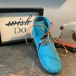 May include: A turquoise blue suede ankle boot with brown laces. The boot is tied with a bow and has a stitched seam around the sole. The boot is sitting on a wooden surface next to a framed sign that says "Do" with the word "wish" crossed out.