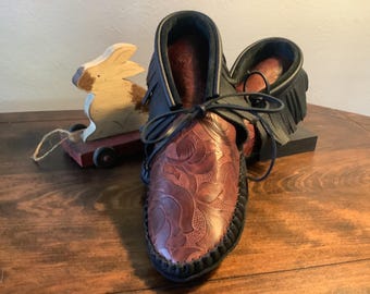 May include: A pair of brown leather moccasins with intricate floral tooling. The moccasins have black leather trim and fringe.