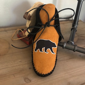 May include: A pair of brown leather moccasins with black stitching and a black bear silhouette embroidered on the front. The moccasins are tied with black laces.