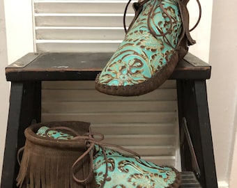 May include: A pair of turquoise leather moccasins with brown suede fringe and laces. The moccasins are sitting on a wooden step stool.