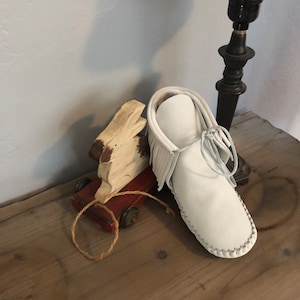 May include: White leather moccasin with fringe detail. The moccasin is sitting on a wooden surface with a toy rabbit and a red wagon.