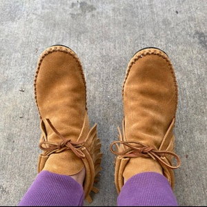 May include: A pair of brown suede ankle boots with fringe detailing and laces. The boots are worn by a person wearing purple pants.