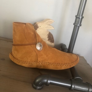 May include: Tan suede moccasin boots with a decorative metal button on the side. The boots have a stitched seam around the sole.