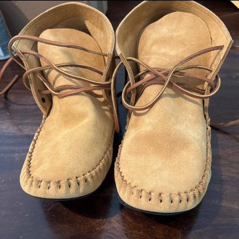 Pu&ograve; includere: Un paio di mocassini in camoscio beige con lacci marroni. I mocassini hanno una cucitura decorativa attorno alla suola e un dettaglio a frangia nella parte superiore.