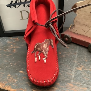 May include: Red leather moccasin with a brown horse embroidered on the front. The moccasin has a brown leather lace and is tied at the top.