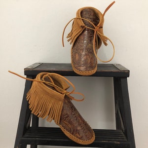 May include: A pair of brown leather moccasins with fringe and a paisley pattern. The moccasins are sitting on a black wooden step stool.
