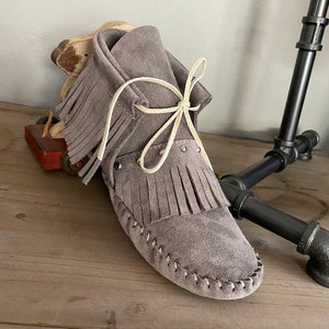 May include: A gray suede moccasin with fringe detailing and a cream-colored lace closure. The shoe features decorative studs and stitched accents along the sole. The moccasin is a slip-on style, perfect for casual wear.