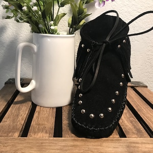 May include: Black suede moccasins with silver studs and fringe. The moccasins are on a wooden surface.