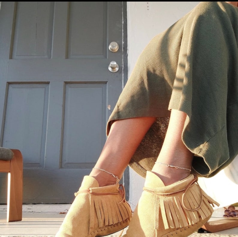 May include: A pair of tan suede ankle boots with fringe detailing and a silver chain ankle bracelet. The boots are worn by a person sitting on a wooden chair.