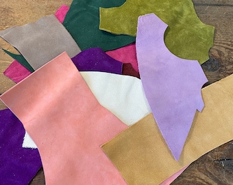May include: Assortment of suede leather scraps in various colors, including purple, pink, green, brown, and white. The scraps are arranged in a pile on a wooden surface.