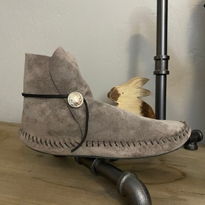 May include: A gray suede moccasin with a black leather strap and a silver button. The moccasin is sitting on a wooden surface.