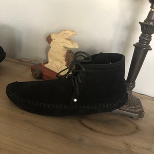 May include: Black suede moccasin boots with a lace-up closure. The boots have a decorative stitch around the sole and a small silver stud on the side.